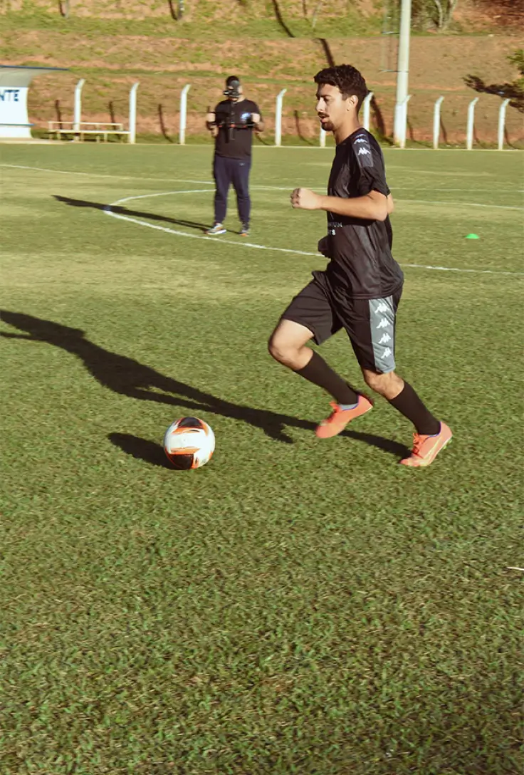 Um jogador de futebol em um uniforme preto e cinza dribla a bola em um campo verde bem cuidado. Outra pessoa está no fundo, registrando o momento.