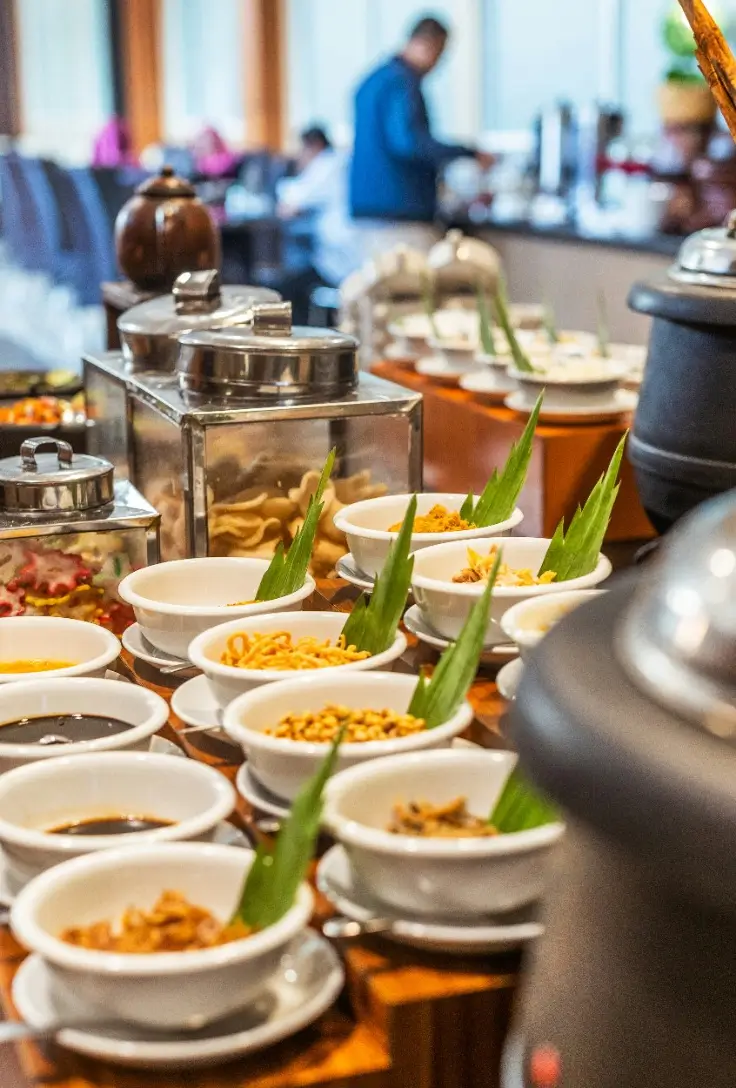 Um buffet de comida com várias opções de pratos, incluindo tigelas de molhos e petiscos, decorado com folhas verdes, em um ambiente interno iluminado.