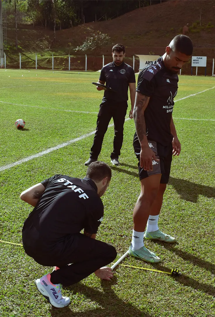 Uma equipe de futebol está em um campo, onde um membro da equipe técnica, identificado pela camiseta com a palavra "STAFF", está medindo a altura do salto de um jogador usando uma fita métrica. Outro membro da equipe observa, segurando um bloco de notas e uma caneta.