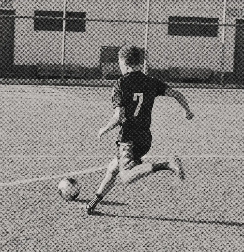 A black and white image of a soccer player with the number 7 on their jersey kicking a ball on a grassy field with a building in the background.